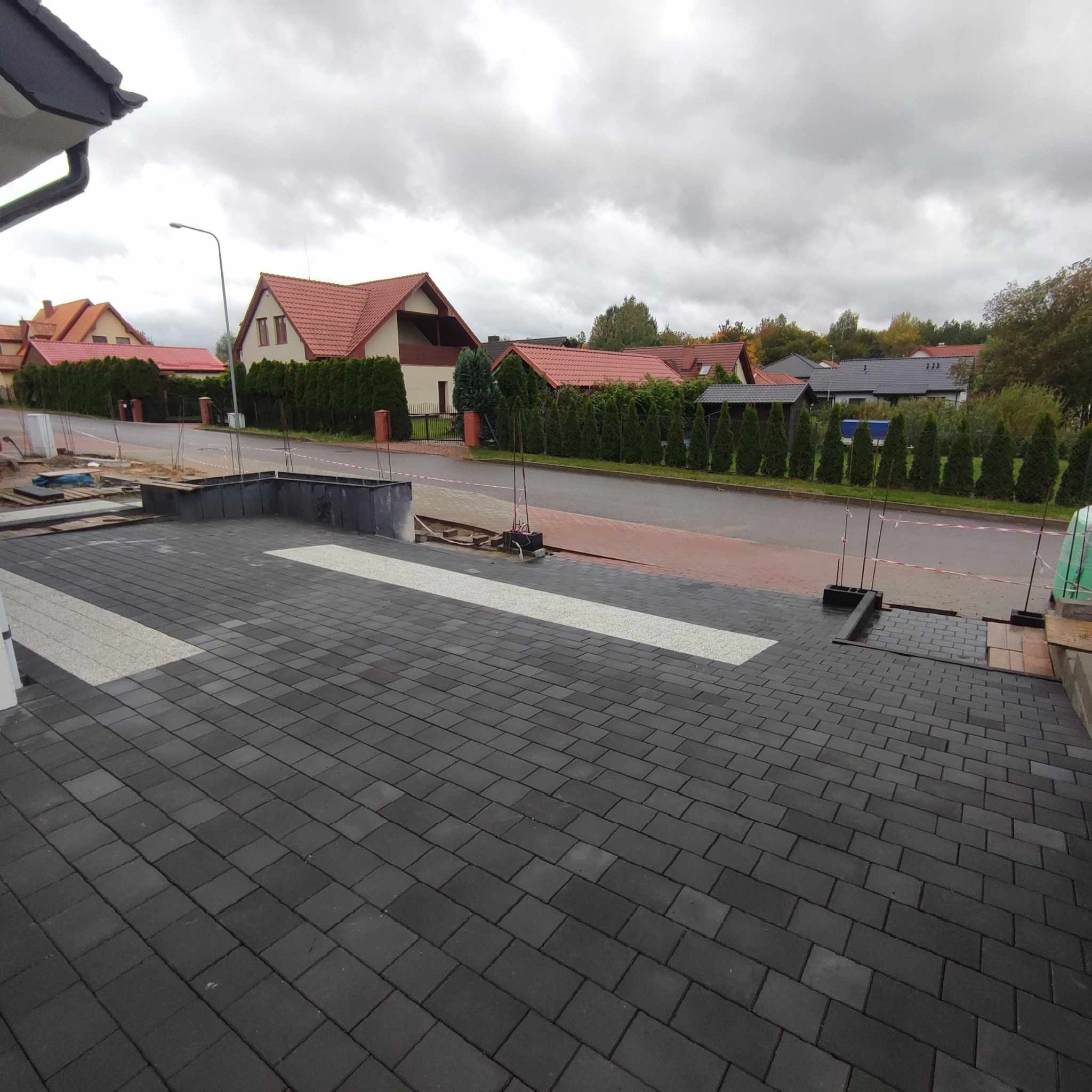 Częściowo ułożona, ciemnoszara kostka brukowa z jasnymi wstawkami na podjeździe, widok na ulicę z domami w tle i pochmurne niebo.