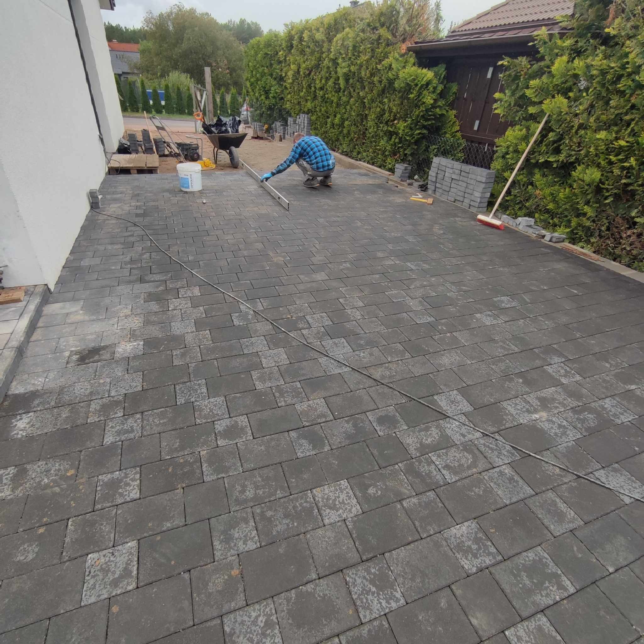 Układanie szarej kostki brukowej na posesji, pracownik w niebieskiej koszuli wyrównuje poziom, w tle taczka z narzędziami i roślinność.