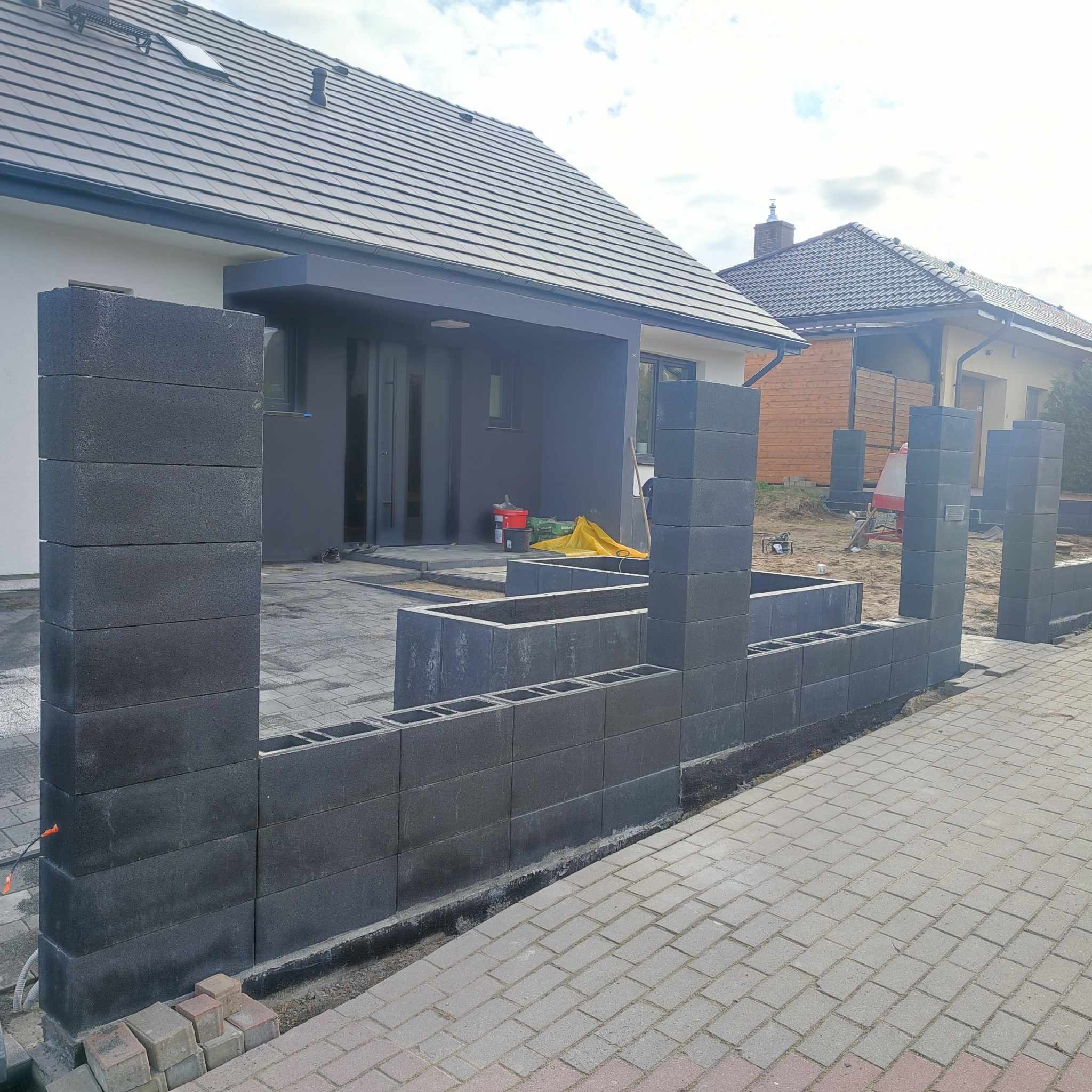 Budowa ogrodzenia z szarych bloczków betonowych przy nowym domu z szarą dachówką, widoczny fragment chodnika z kostki brukowej i narzędzia budowlane w tle.