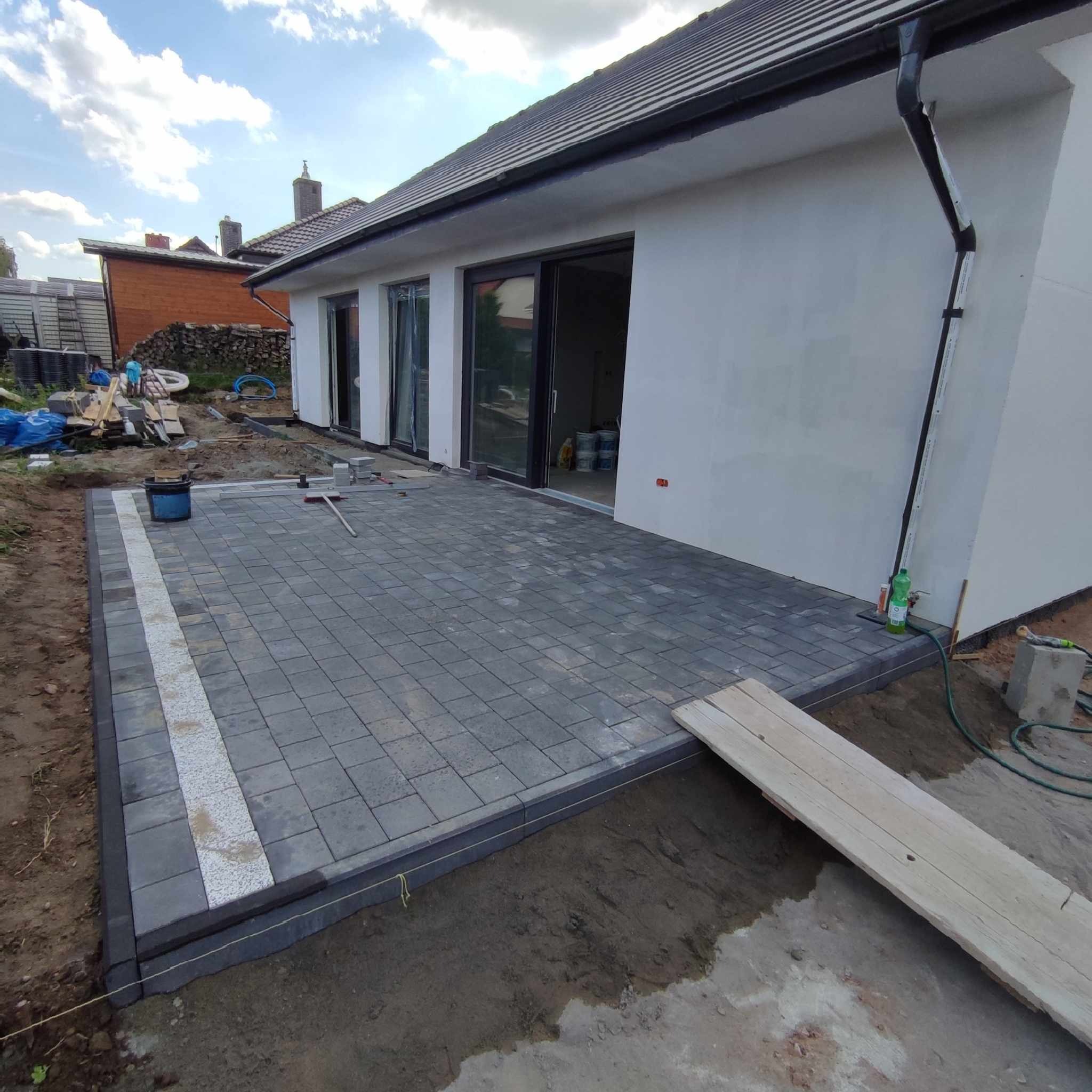 Nowo ułożony taras z szarej kostki brukowej z białym pasem dekoracyjnym, widok na dom z dużymi oknami przesuwnymi, teren wokół w trakcie prac ziemnych.