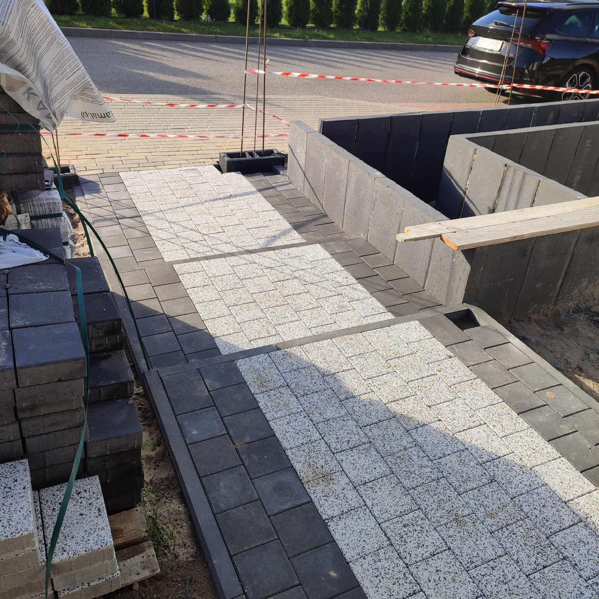Wykładanie jasnej kostki brukowej w Piła, obrzeża z ciemnej kostki, w tle zaparkowany samochód i zabezpieczenia budowlane.