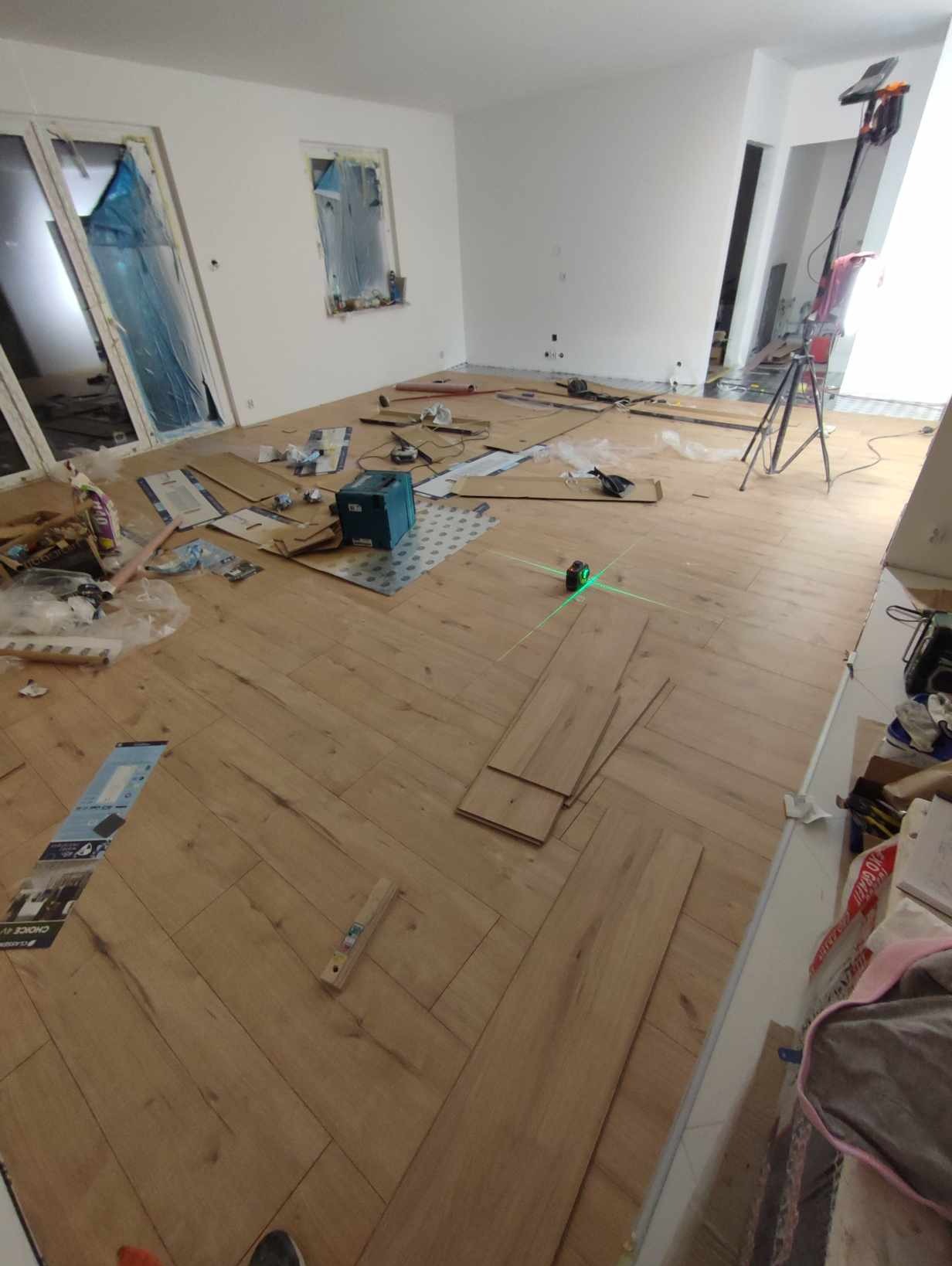 Układanie parkietu w jodełkę w jasnym pomieszczeniu, widoczny laser krzyżowy, opakowania po panelach i narzędzia pracy.