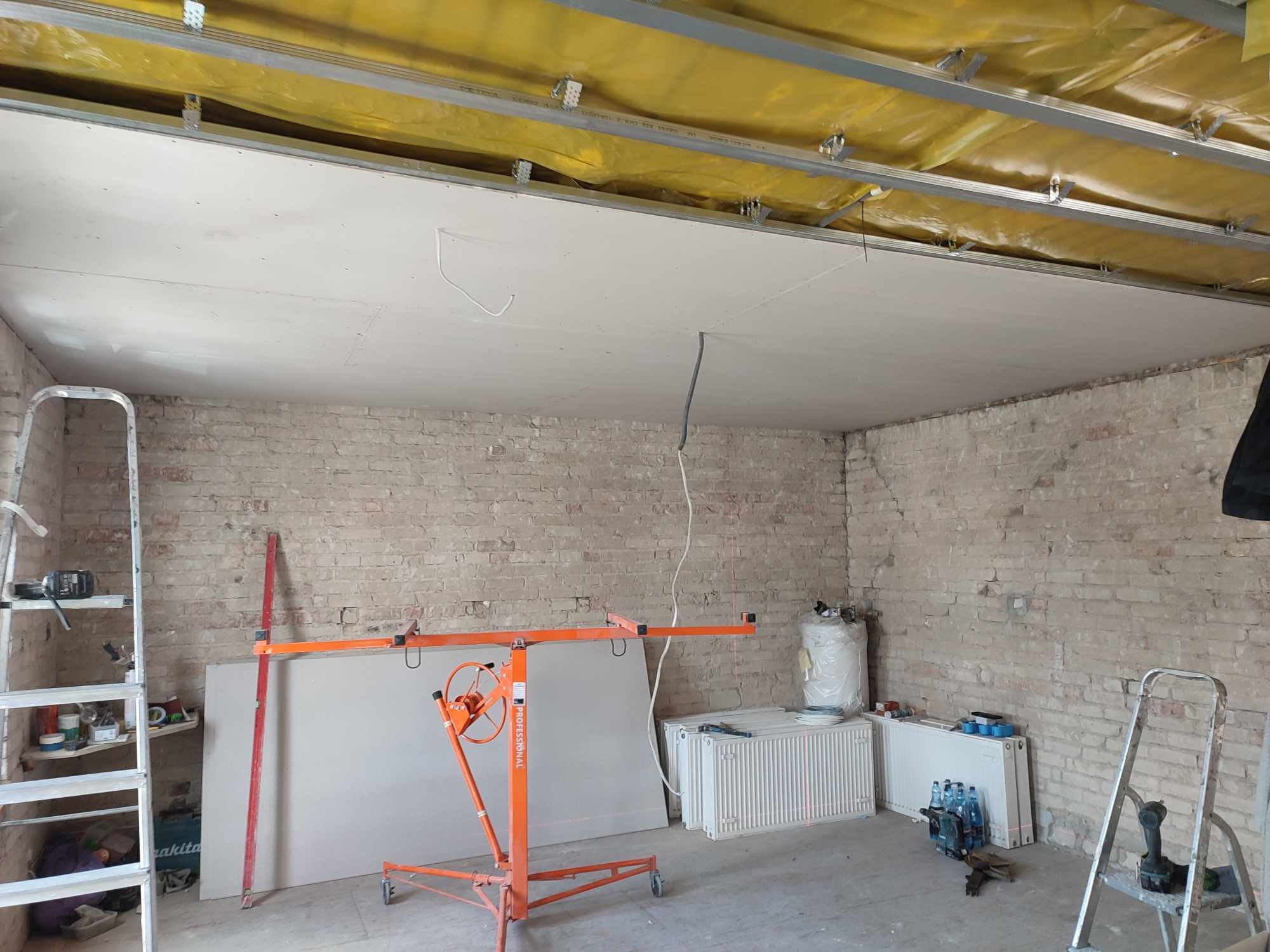 Montaż sufitu podwieszanego z płyt gipsowo-kartonowych, widoczny stelaż, ocieplenie wełną mineralną i podnośnik do płyt na tle surowej ceglanej ściany.