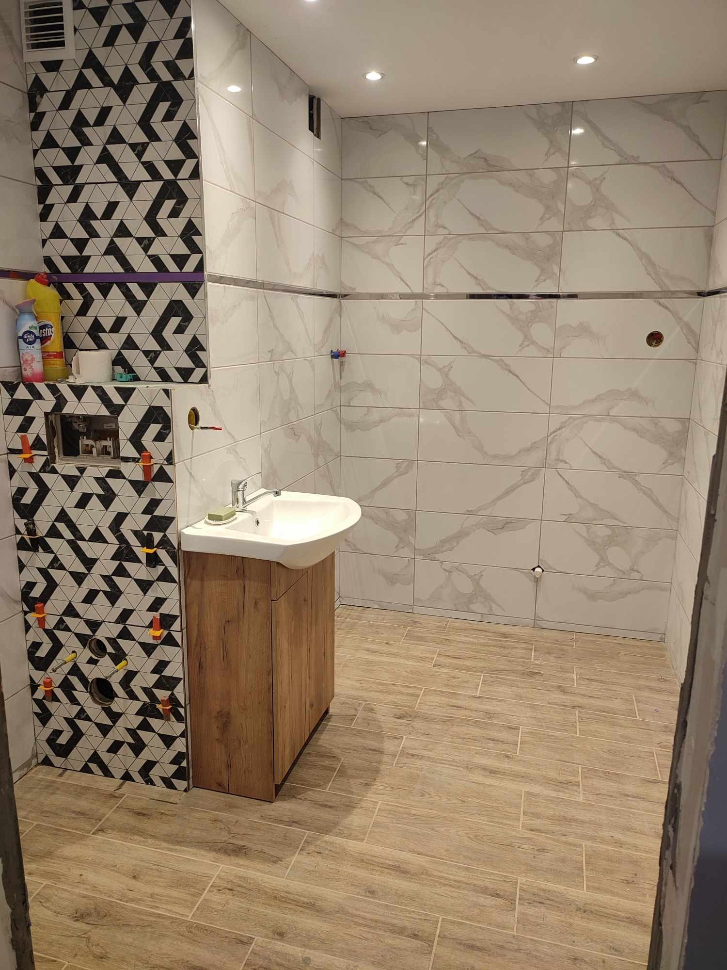 Łazienka w trakcie prac wykończeniowych: białe płytki imitujące marmur na ścianach, drewnopodobna podłoga, umywalka z szafką, fragment ściany z czarno-białą mozaiką, widoczne krzyżyki dystansowe.