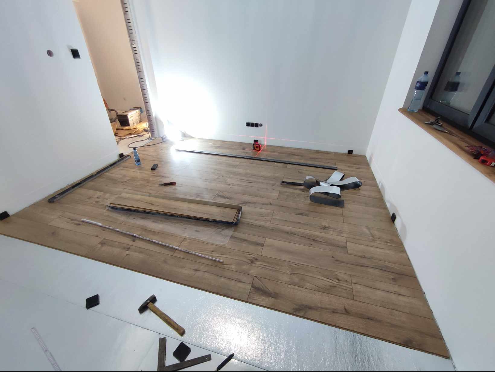 Podłoga z paneli w trakcie układania, widoczna folia izolacyjna, listwy, narzędzia i poziomica laserowa.