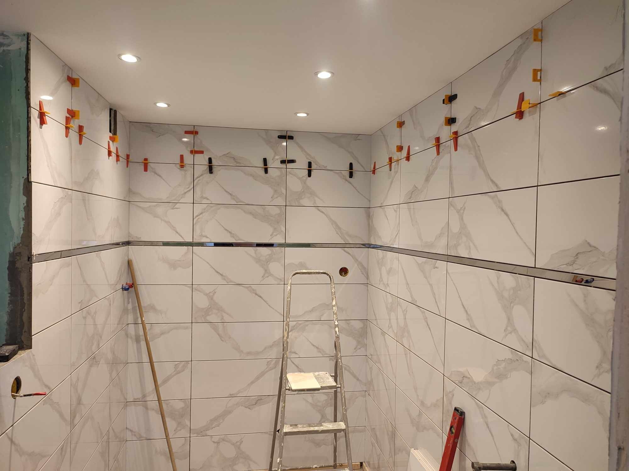 Ściany łazienki wyłożone białymi płytkami imitującymi marmur, z użyciem klipsów poziomujących, drabina w centrum kadru.