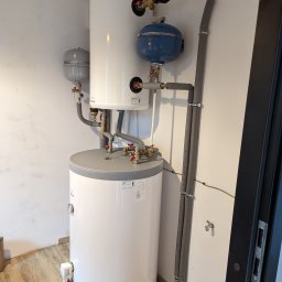 STANTECH PIOTR STANKIEWICZ - Kaskadowe połączenie dwóch białych zbiorników akumulacyjnych ciepłej wody z osprzętem, widoczne zawory, manometry, pompy obiegowe i izolowane rury w pomieszczeniu technicznym.