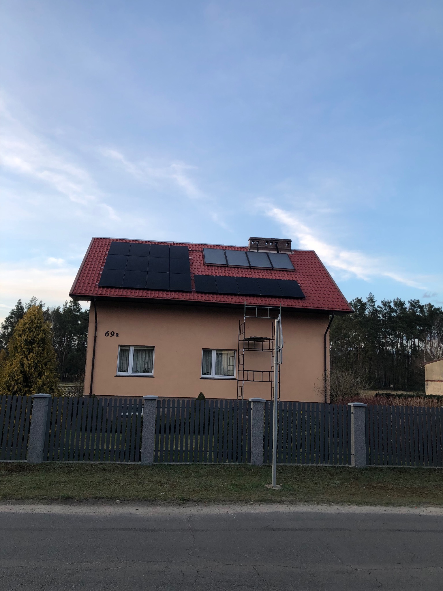 Dom jednorodzinny z panelami słonecznymi na czerwonym dachu, widoczne rusztowanie i numer domu 69a.