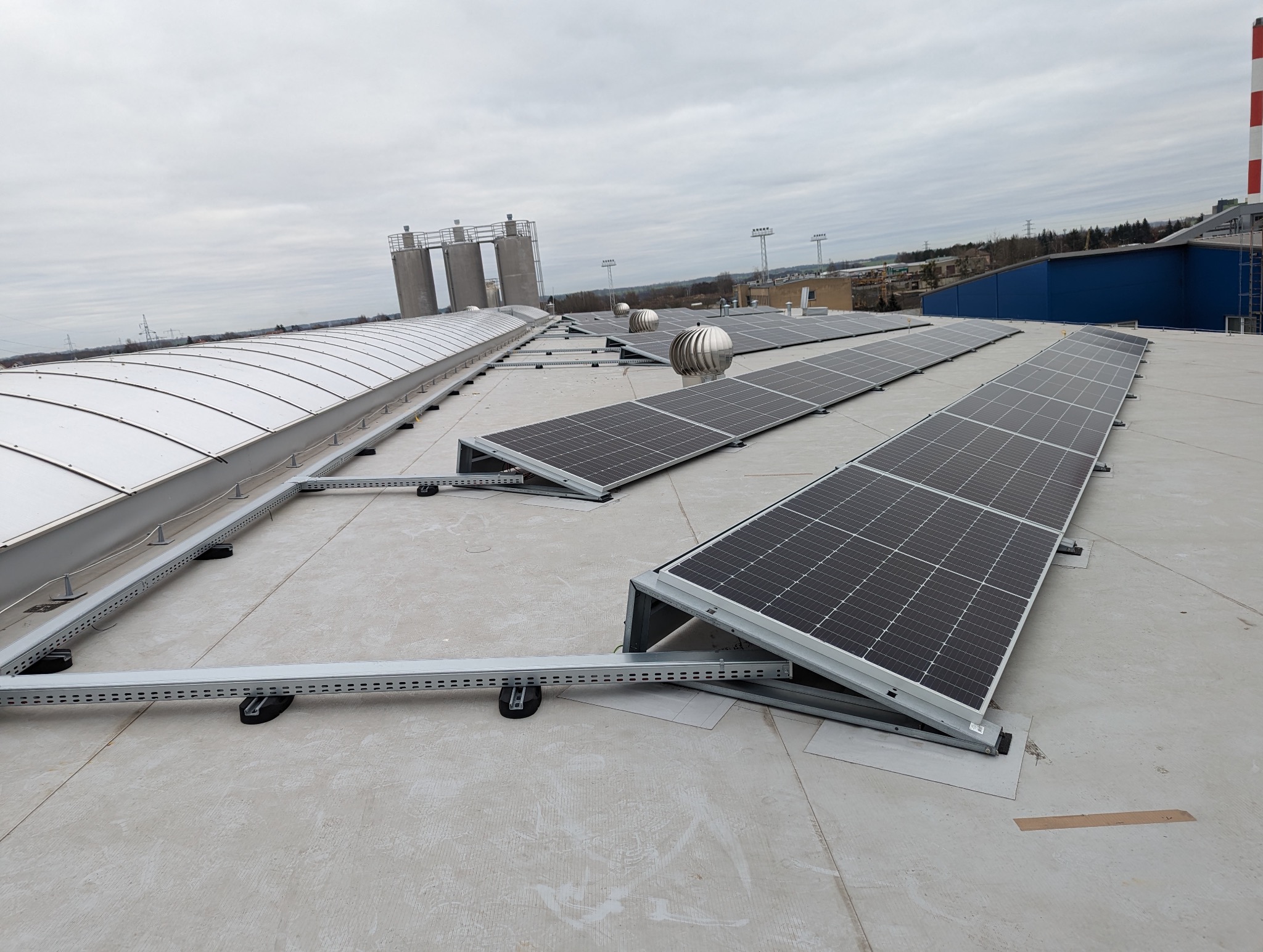 Instalacja fotowoltaiczna na dachu przemysłowym z widocznymi panelami, systemem montażowym i elementami wentylacji dachowej w pochmurny dzień.