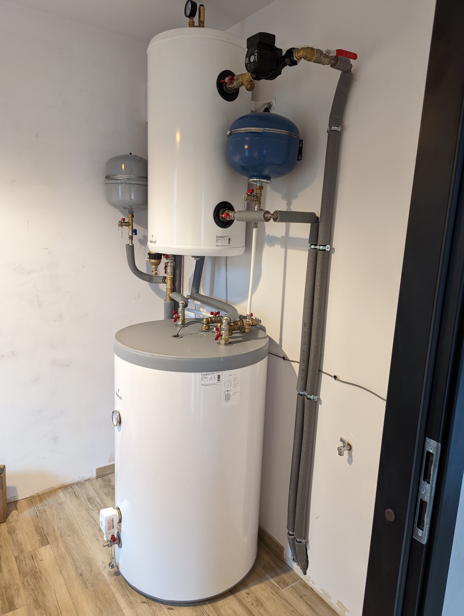 Kaskadowe połączenie dwóch białych zbiorników akumulacyjnych ciepłej wody z osprzętem, widoczne zawory, manometry, pompy obiegowe i izolowane rury w pomieszczeniu technicznym.