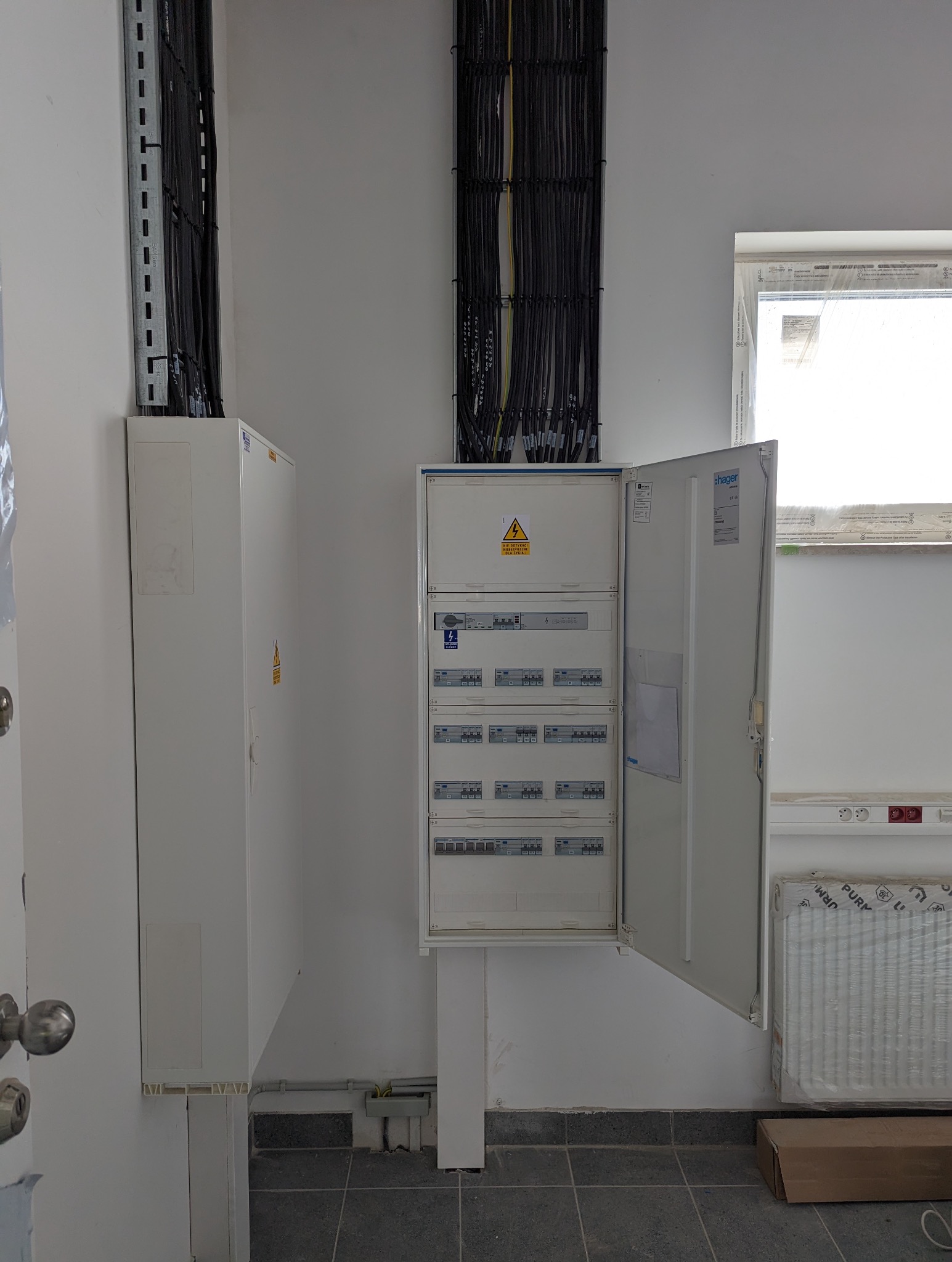 Otwarta skrzynka rozdzielcza Hager z widocznymi bezpiecznikami i przewodami, montowana na ścianie w nowym budynku, z przewodami prowadzonymi w korytach kablowych.