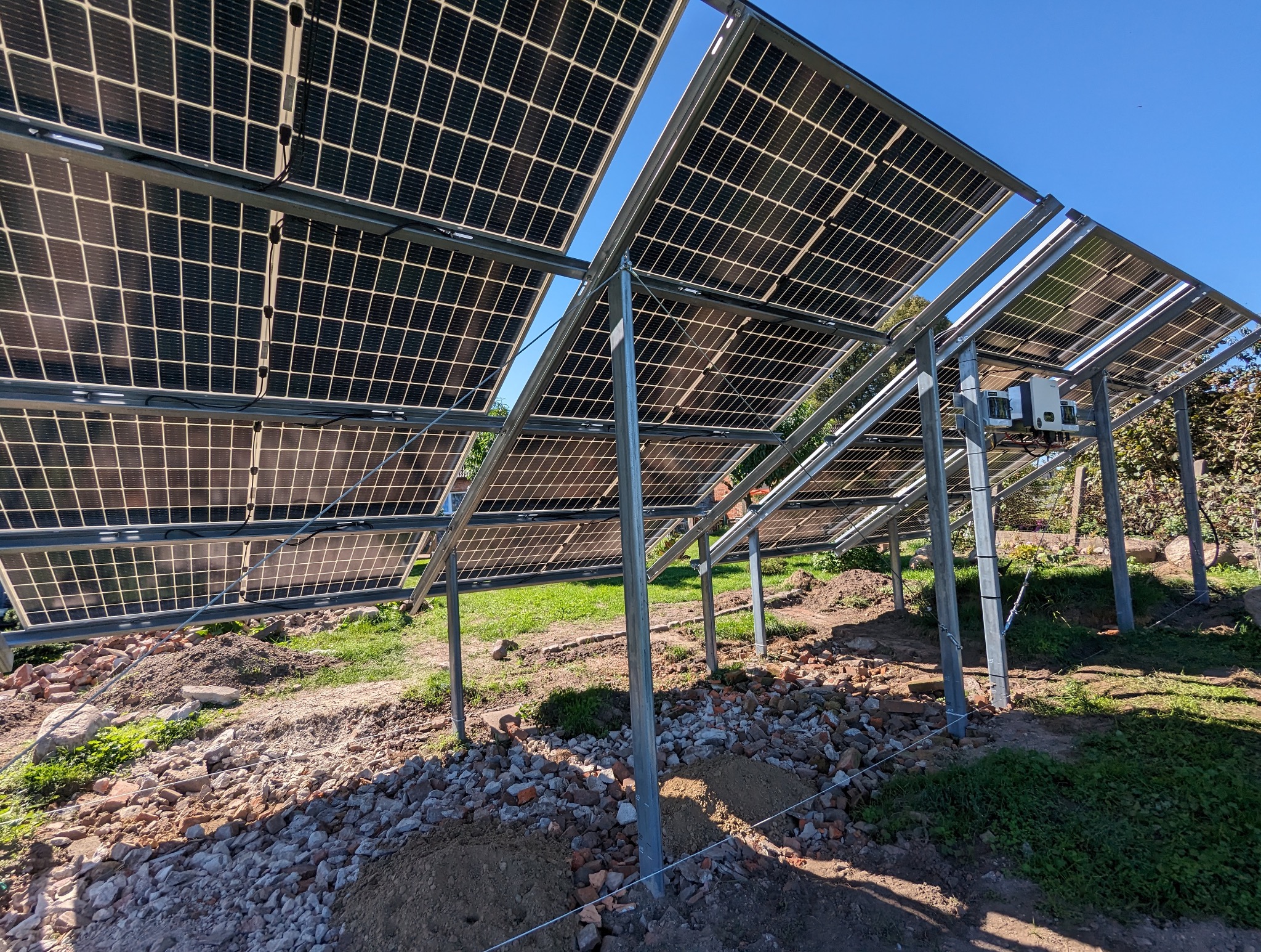 Widok od dołu na konstrukcję wsporczą paneli fotowoltaicznych z zamontowanymi panelami, widoczne inwertery, teren wokół w trakcie prac ziemnych, niebo w tle.