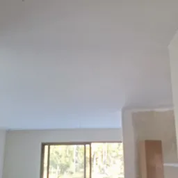 Wnętrze pokoju po szpachlowaniu, widoczny gładki, biały sufit i ściany, z oknem w tle i fragmentem ściany przygotowanej do malowania.