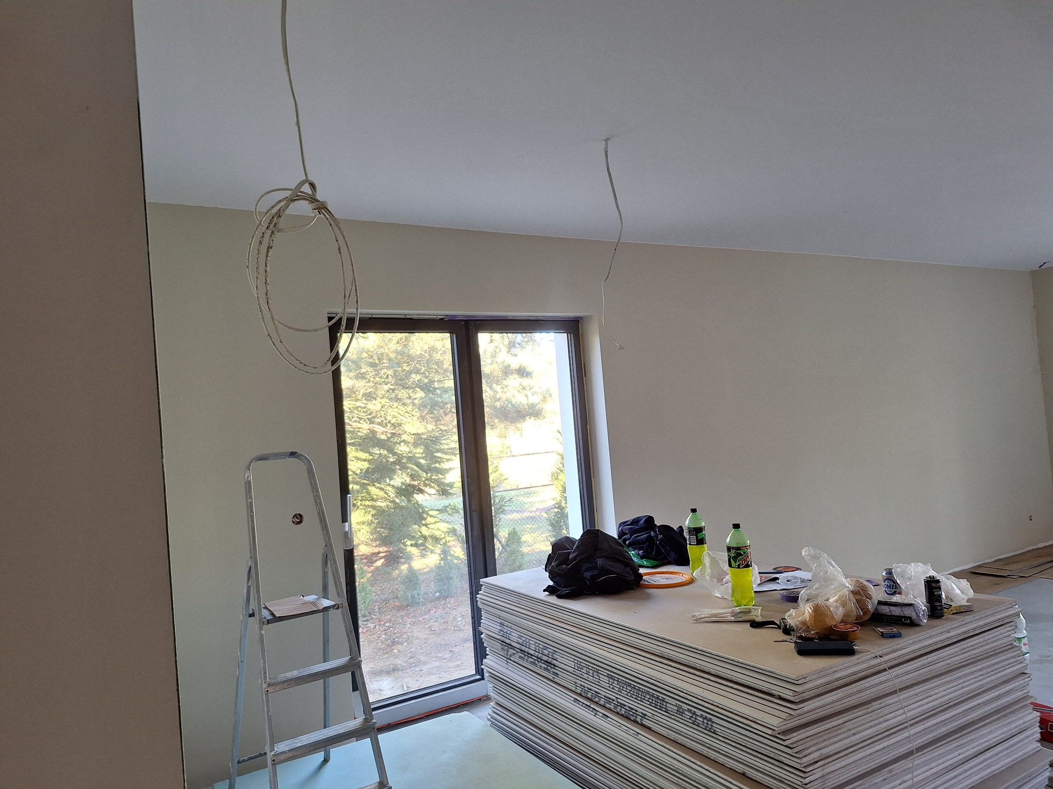 Stos płyt gipsowo-kartonowych w surowym wnętrzu, obok okna z widokiem na zieleń, drabina aluminiowa i wiszące kable elektryczne.