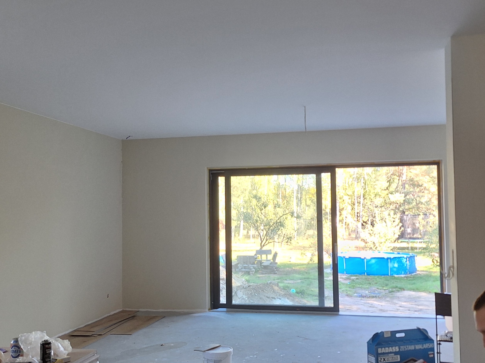 Wnętrze pokoju w Łodzi po świeżym malowaniu ścian na jasny odcień beżu, z widocznymi listwami przypodłogowymi i dużym oknem przesuwnym z widokiem na ogród z basenem.