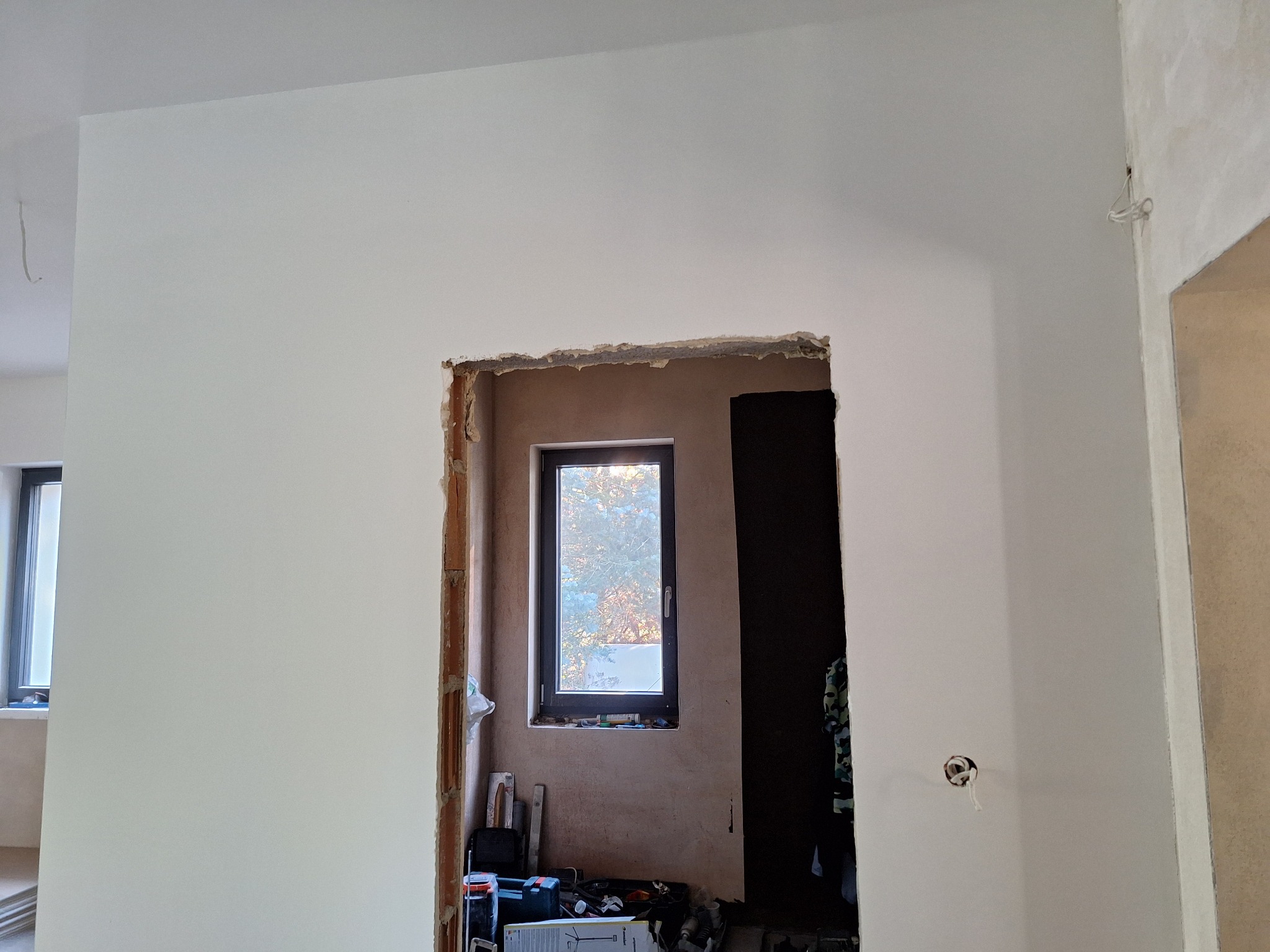 Wnętrze w trakcie remontu: ściany częściowo pomalowane na biało, widoczne niedokończone obramowanie otworu drzwiowego z cegieł, okno z widokiem na zewnątrz, narzędzia i materiały budowlane...