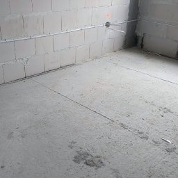 Dominik Gołębiewski - Surowy stan: szara ściana z betonu komórkowego z widoczną, poziomą instalacją elektryczną w białej rurce, surowy beton na podłodze. Wnętrze w trakcie prac wykończeniowych.