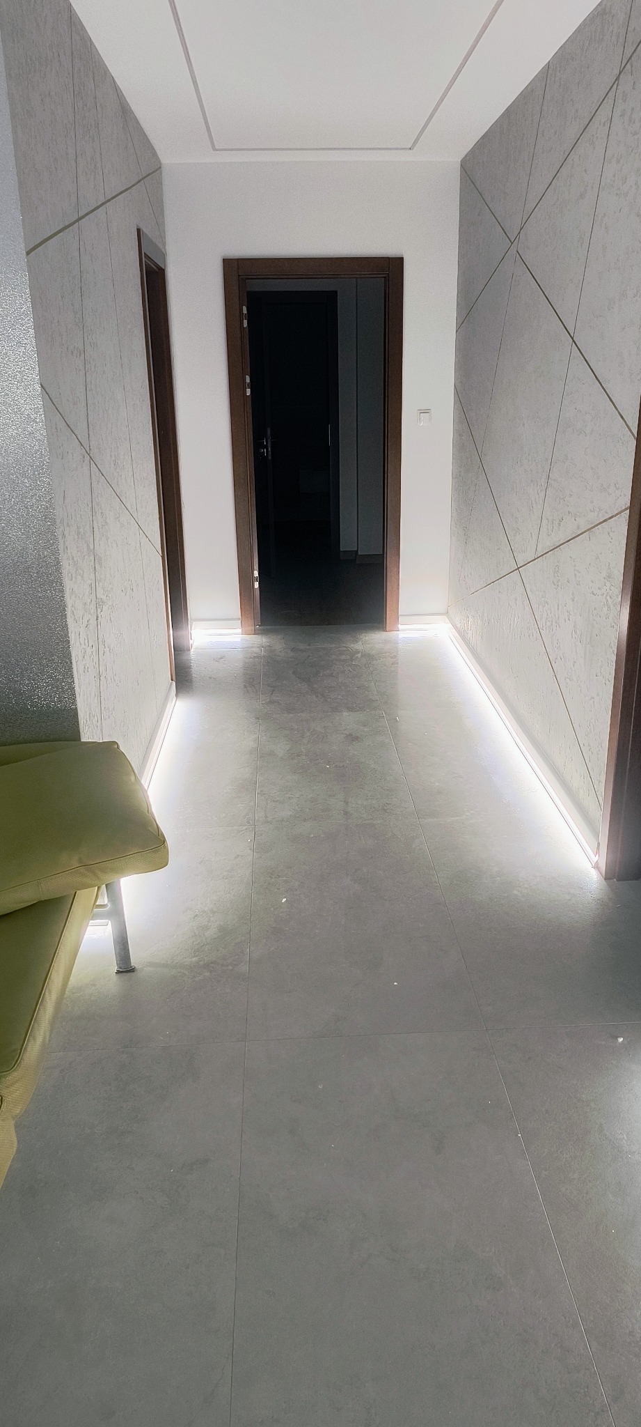 Nowoczesny korytarz z podświetleniem LED wzdłuż ścian, szare płytki podłogowe i dekoracyjne panele ścienne z geometrycznym wzorem. Widoczne drzwi wewnętrzne.