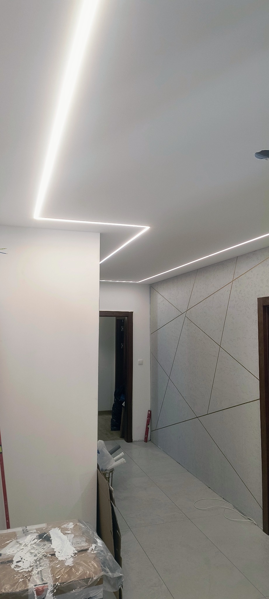 Nowoczesne oświetlenie LED w korytarzu z geometryczną ścianą. Białe, liniowe lampy sufitowe tworzą unikalny wzór świetlny. Widoczne elementy wykończenia wnętrz.