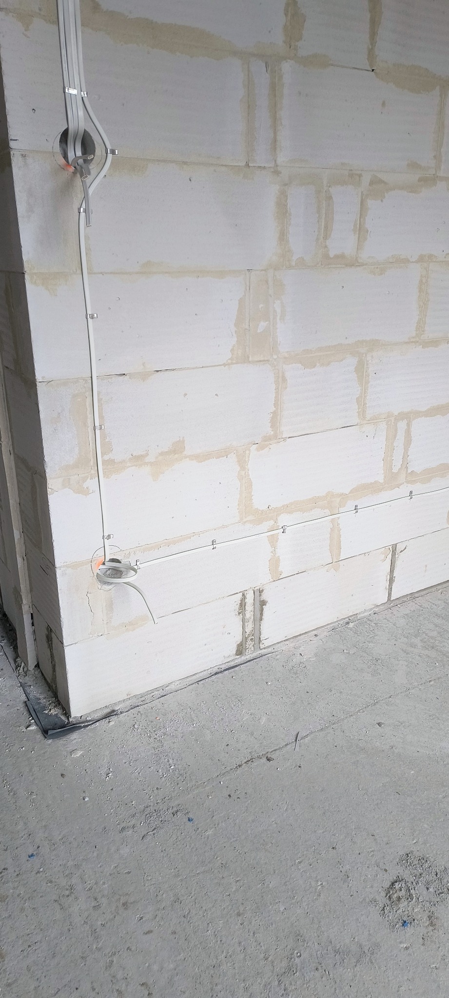 Biała ściana z betonu komórkowego z poprowadzonymi przewodami elektrycznymi, zamocowanymi klamrami. Widoczne puszki instalacyjne. Surowy stan wykończenia.