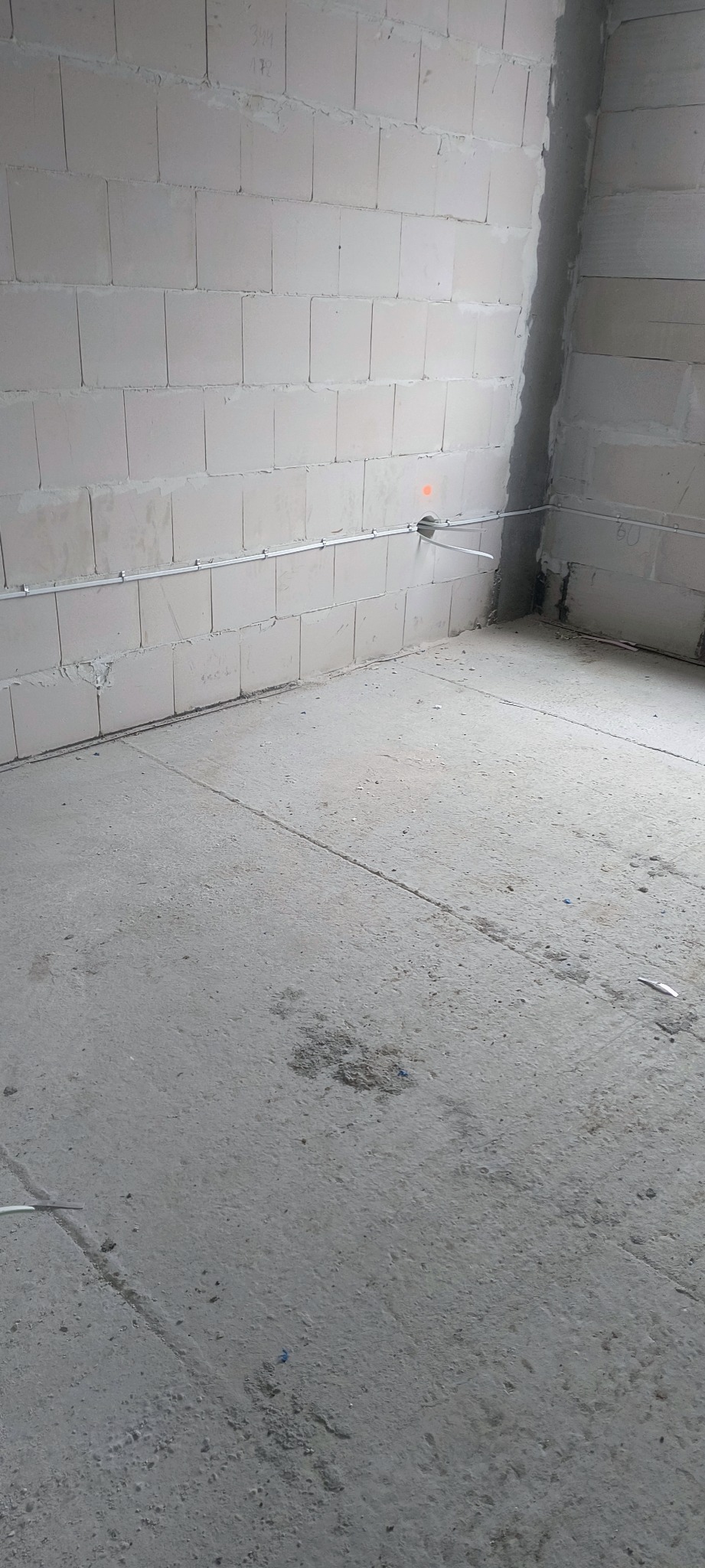Surowy stan: szara ściana z betonu komórkowego z widoczną, poziomą instalacją elektryczną w białej rurce, surowy beton na podłodze. Wnętrze w trakcie prac wykończeniowych.