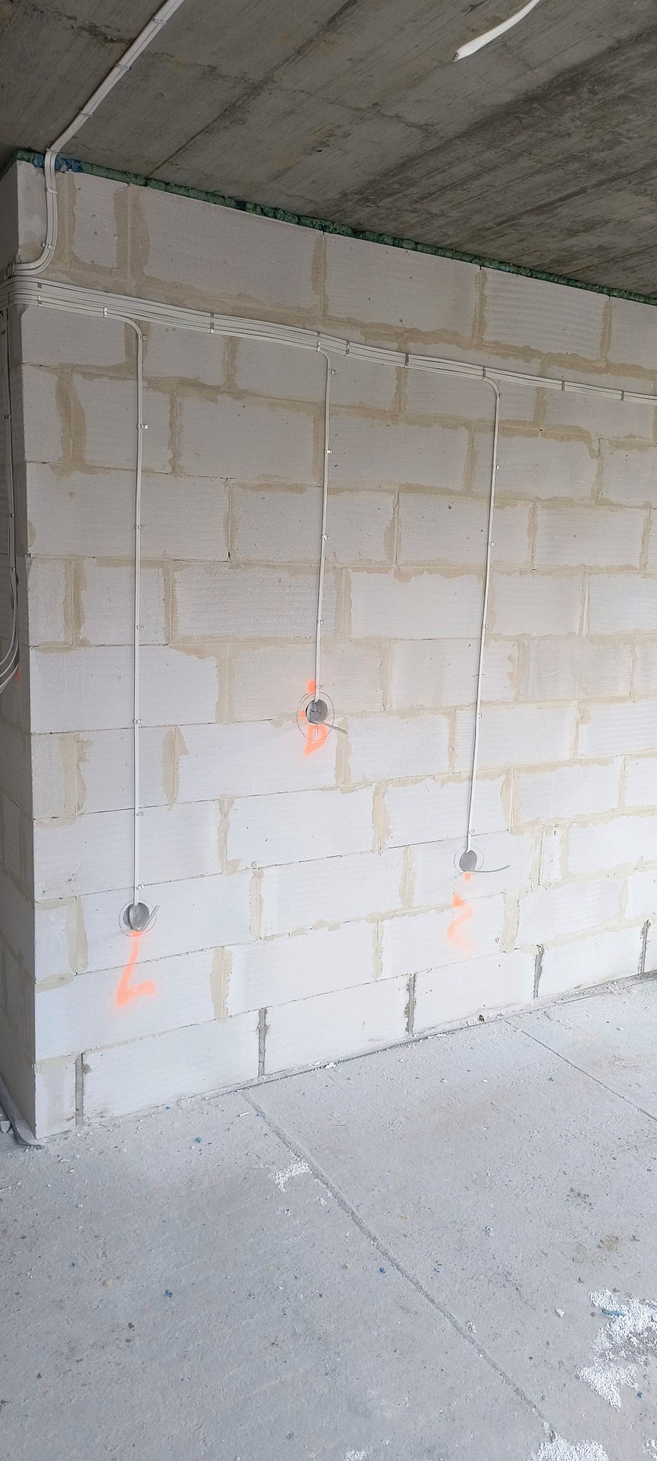 Ściana z betonu komórkowego z poprowadzonymi przewodami elektrycznymi w białych listwach. Widoczne otwory pod gniazdka oznaczone pomarańczową farbą.