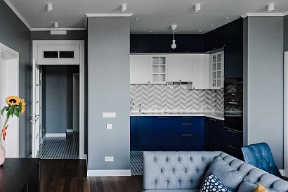 Nowoczesna kuchnia z granatowo-białymi szafkami, szarą ścianą i chevronowym backsplashiem, widok z salonu z szarą sofą.