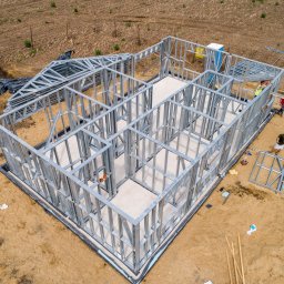 Stelnest - Domy Szkieletowe ze stali - Widok z góry na plac budowy domu szkieletowego z metalową konstrukcją, widoczni pracownicy w trakcie montażu, teren wokół w stanie surowym.