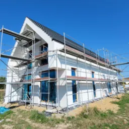 Wznoszony, dwukondygnacyjny dom z białymi ścianami i ciemną dachówką, zabezpieczone okna folią, otoczony rusztowaniem, na tle błękitnego nieba.