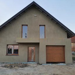 Stelnest - Domy Szkieletowe ze stali - Nowoczesny dom jednorodzinny z garażem w trakcie wykończenia elewacji zewnętrznej, widoczne okna z drewnianymi ramami, brama garażowa i częściowo otynkowana ściana, w tle budynek gospodarczy...