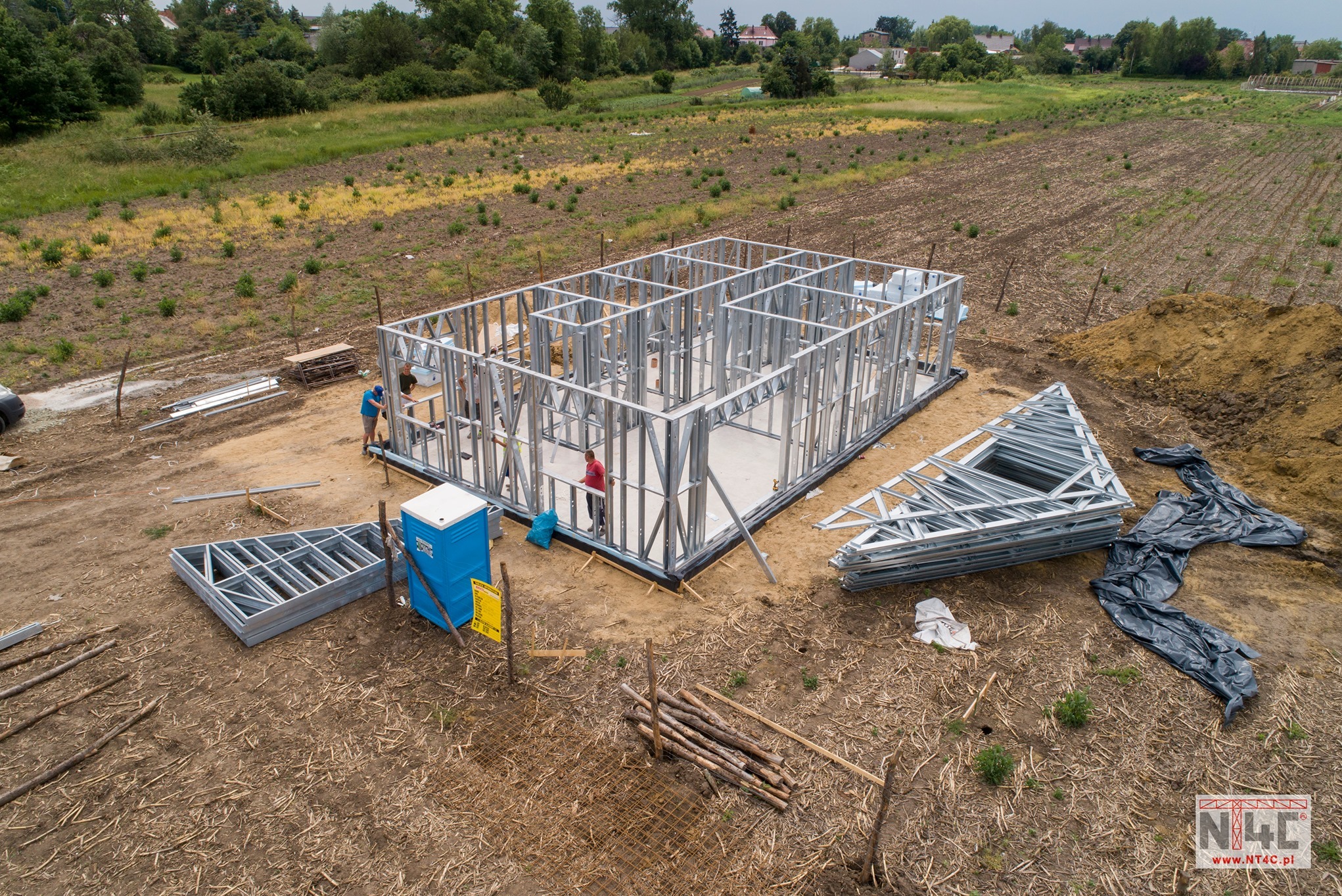 Montaż metalowego szkieletu domu na fundamencie, widok z góry, z pracownikami w tle i przenośną toaletą na placu budowy.