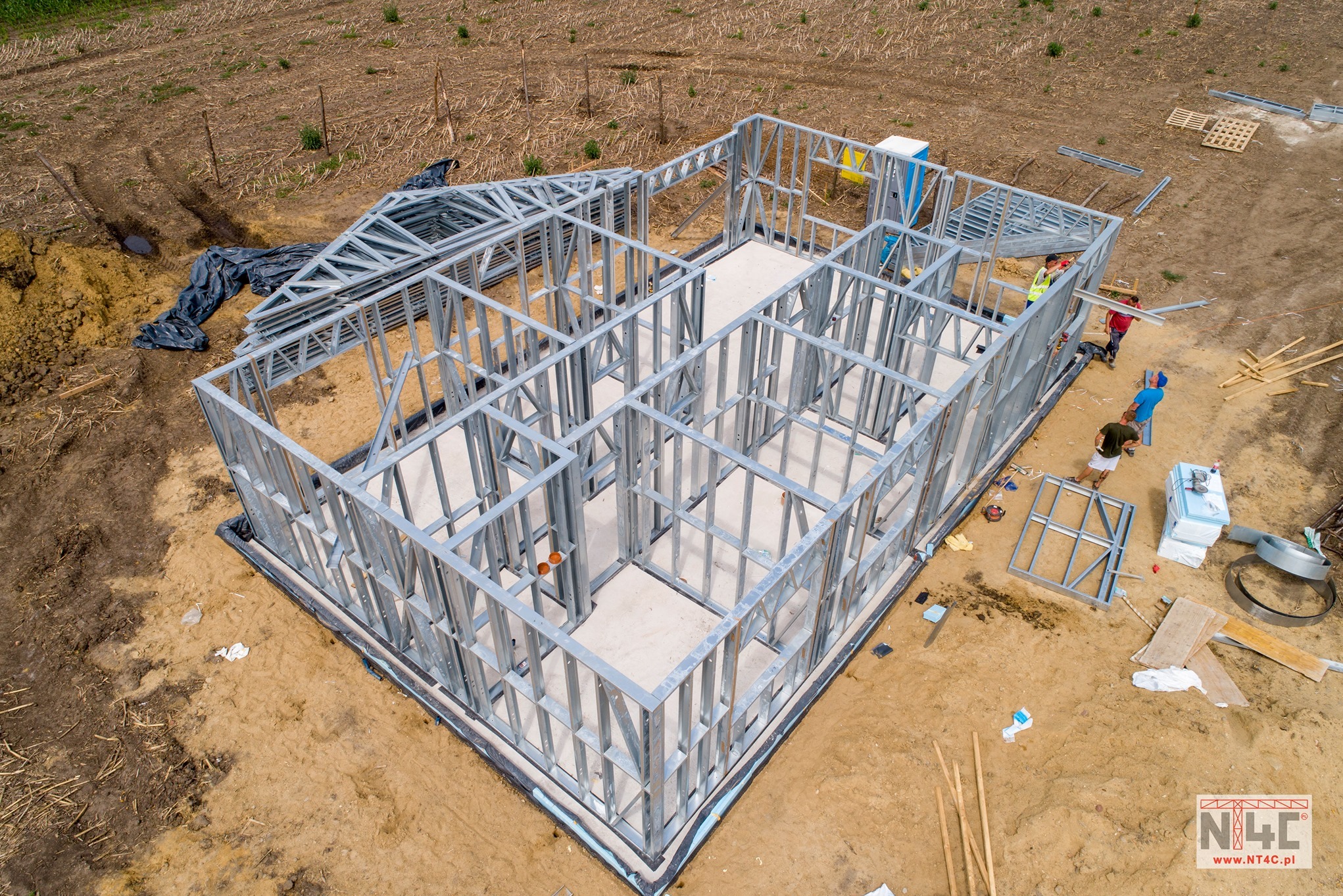 Widok z góry na plac budowy domu szkieletowego z metalową konstrukcją, widoczni pracownicy w trakcie montażu, teren wokół w stanie surowym.