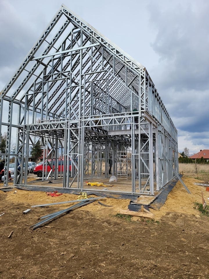 Konstrukcja domu ze stalowych profili na etapie montażu, widoczne narzędzia i fragmenty profili na placu budowy, pochmurne niebo w tle.
