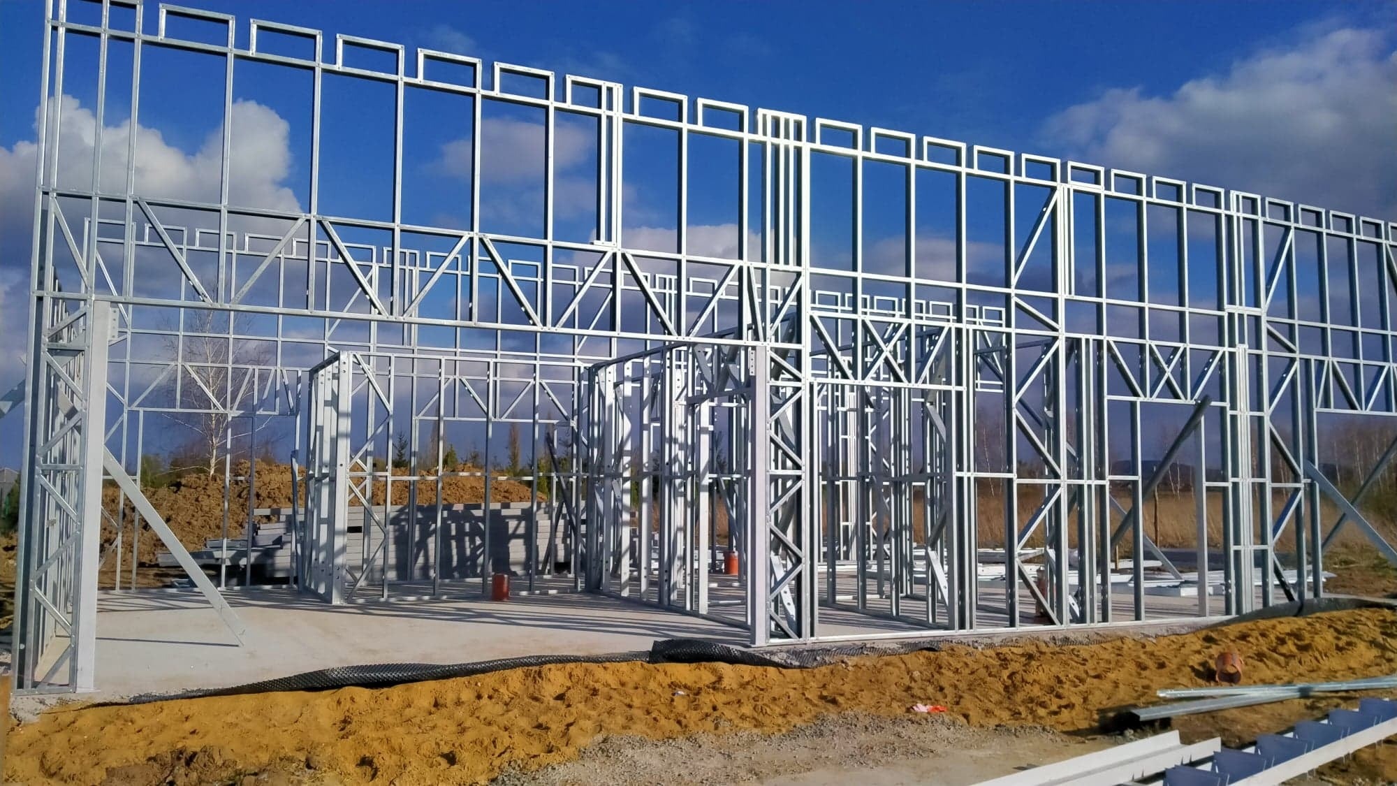 Konstrukcja stalowa domu w trakcie budowy, widoczne elementy szkieletu na tle błękitnego nieba z chmurami.