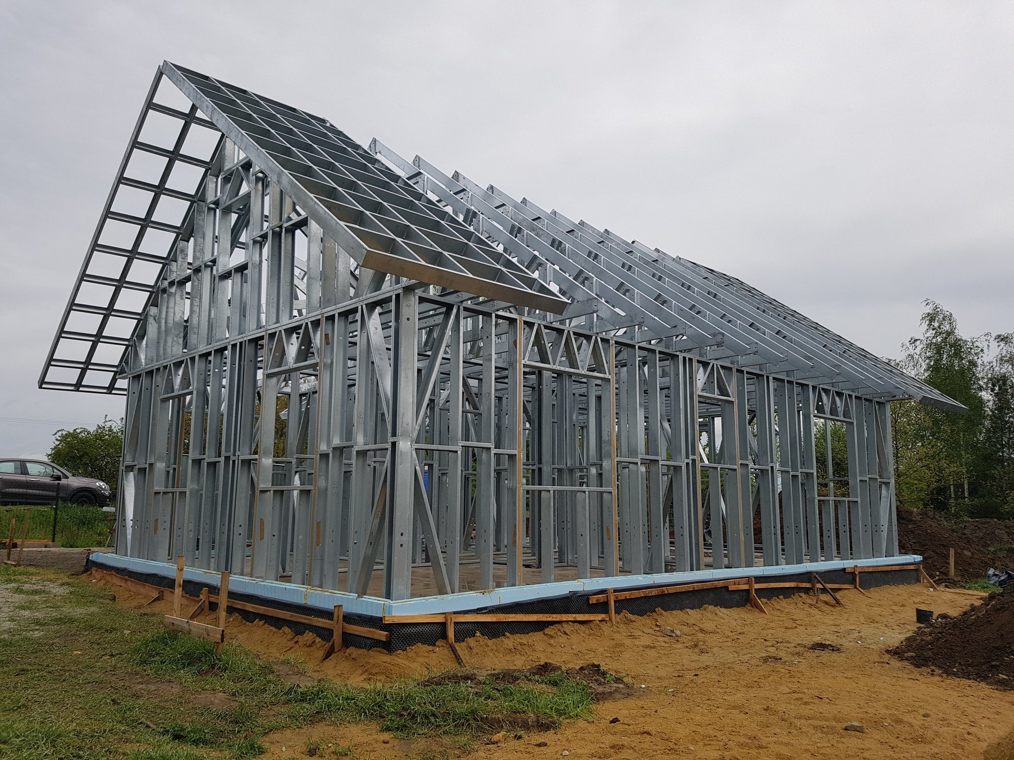 Metalowa konstrukcja domu w trakcie budowy, widoczne stalowe profile ścian i dachu na fundamencie z izolacją, w tle zieleń i samochód.
