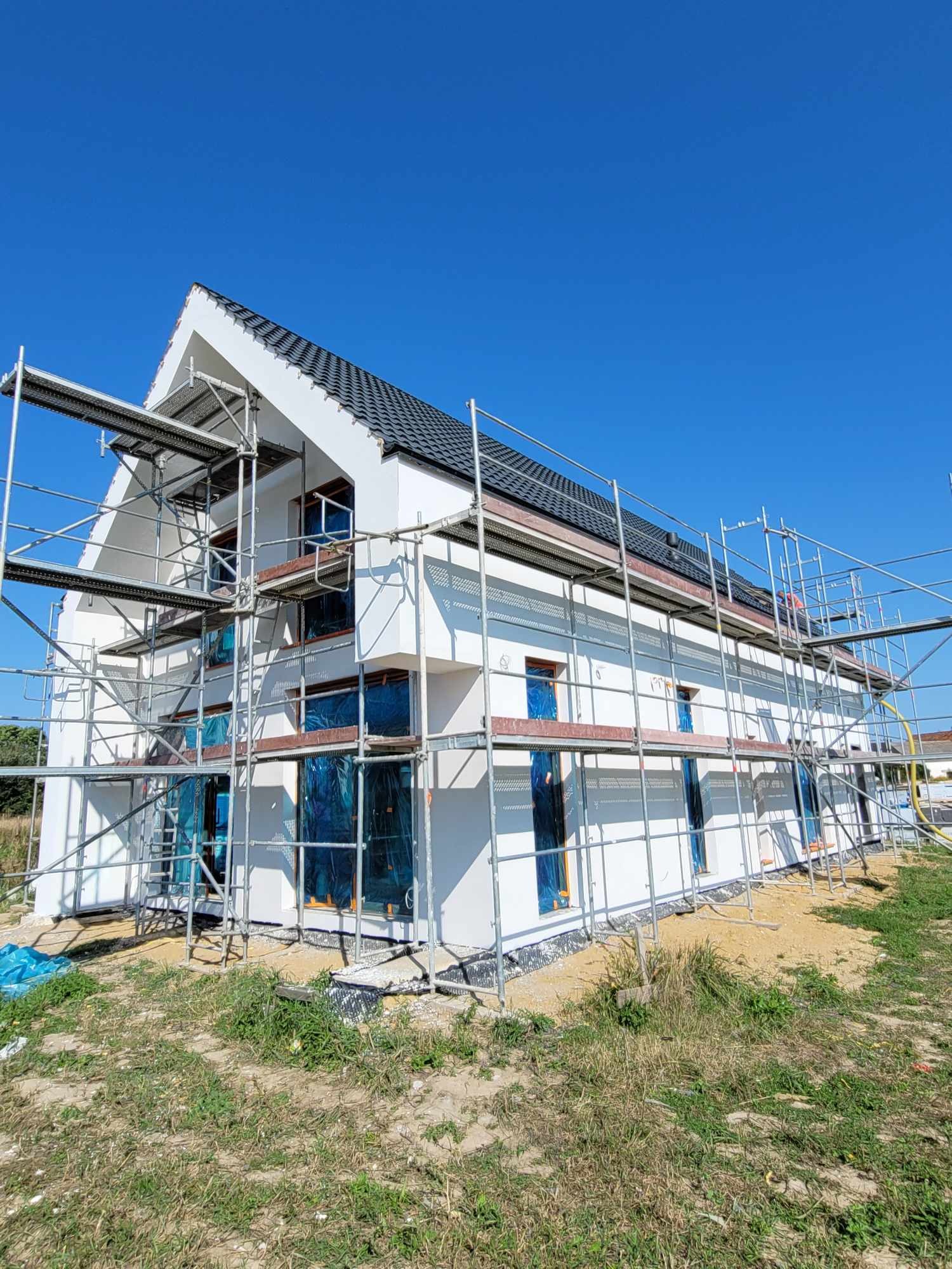 Wznoszony, dwukondygnacyjny dom z białymi ścianami i ciemną dachówką, zabezpieczone okna folią, otoczony rusztowaniem, na tle błękitnego nieba.