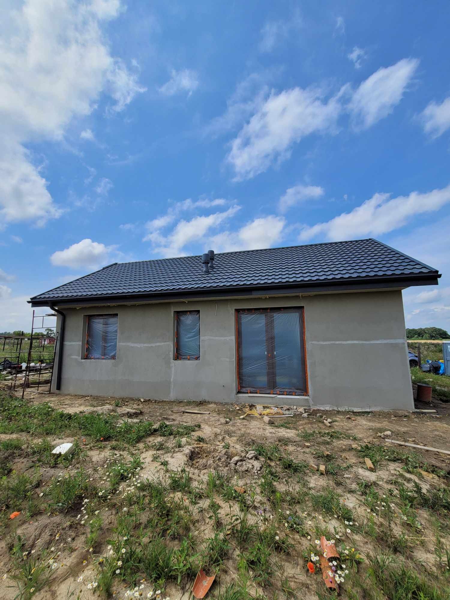 Surowy dom jednorodzinny z szarą elewacją i grafitowym dachem, okna zabezpieczone folią, widok na plac budowy z niezagospodarowanym terenem.