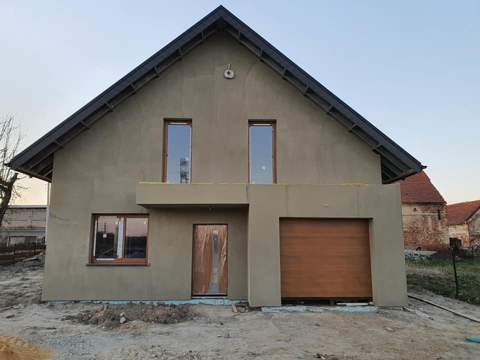 Nowoczesny dom jednorodzinny z garażem w trakcie wykończenia elewacji zewnętrznej, widoczne okna z drewnianymi ramami, brama garażowa i częściowo otynkowana ściana, w tle budynek gospodarczy...