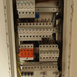 Otwarta skrzynka elektryczna z bezpiecznikami, przewodami i zabezpieczeniami przepięciowymi marki Eaton i Simtec, widok z góry.