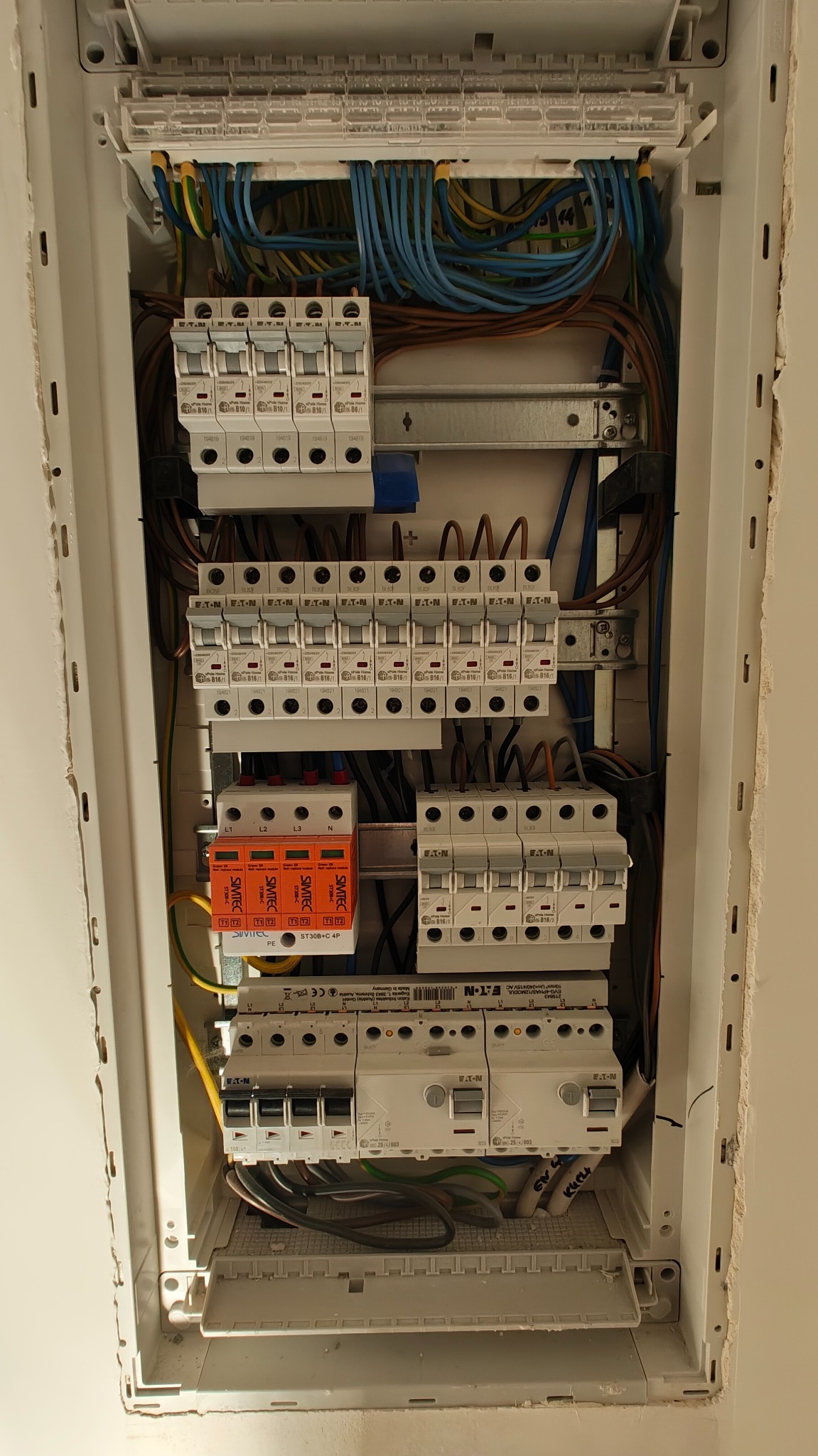 Otwarta skrzynka elektryczna z bezpiecznikami, przewodami i zabezpieczeniami przepięciowymi marki Eaton i Simtec, widok z góry.