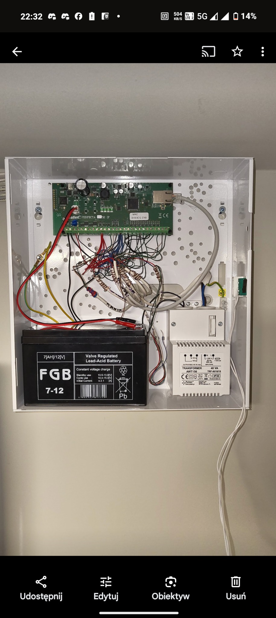 Otwarta biała skrzynka z centralą alarmową Satel Perfecta 32 IP, akumulatorem FGB 7-12 i transformatorem Pulsar AWT150, widoczne liczne przewody i złącza.