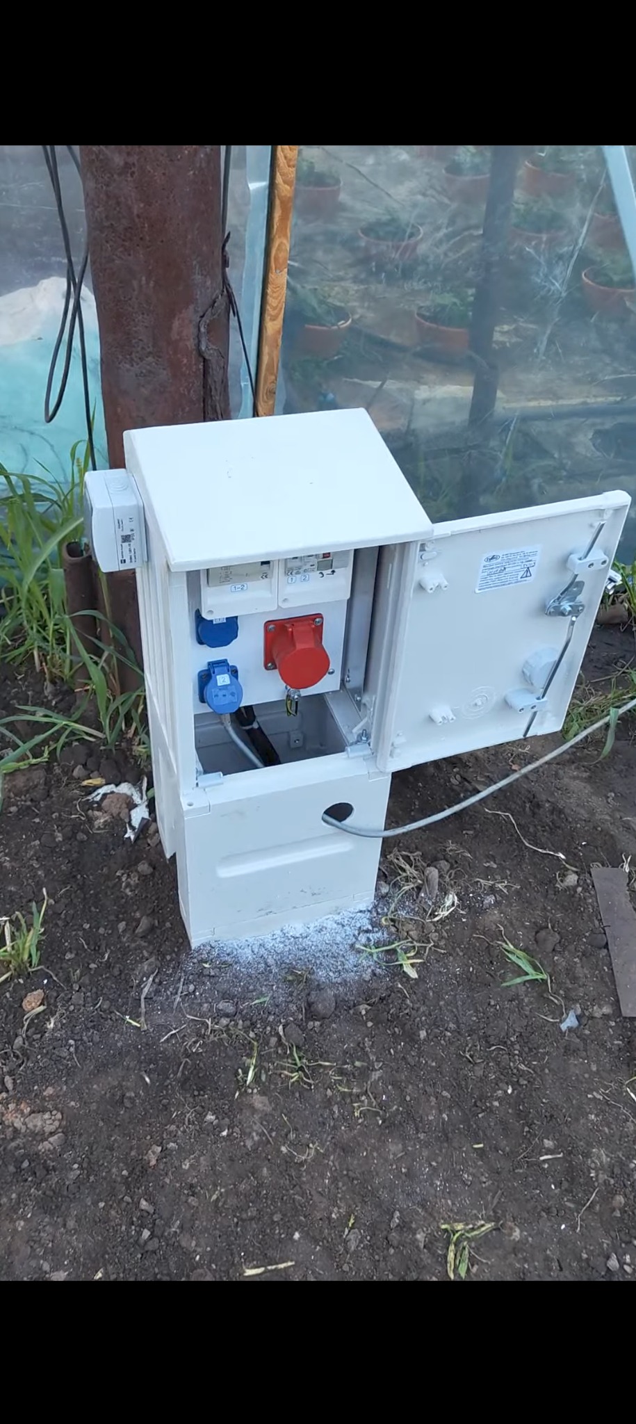 Otwarta skrzynka elektryczna z gniazdami siłowymi (czerwone i niebieskie) i wyłącznikami, zamontowana na zewnątrz, na ziemi, obok szklarni. Widoczne przewody wchodzące do skrzynki.