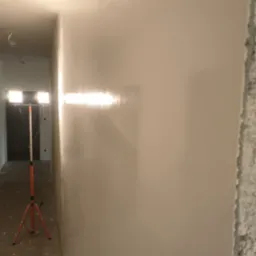 Świeżo nałożona, gładka ściana oświetlona lampą roboczą na trójnogu; widoczny surowy narożnik ściany i przewody pod sufitem.