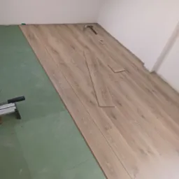 Proces układania paneli podłogowych w pomieszczeniu, widoczna zielona podkładka izolacyjna, częściowo ułożone panele imitujące drewno, narzędzia do cięcia paneli i młotek w tle.