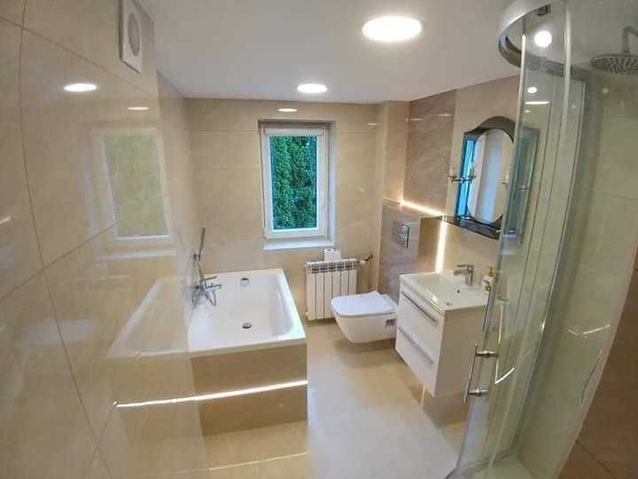 Jasna łazienka z beżowymi płytkami, kabiną prysznicową z zaokrąglonymi drzwiami, wanną z hydromasażem i oknem z widokiem na zieleń.