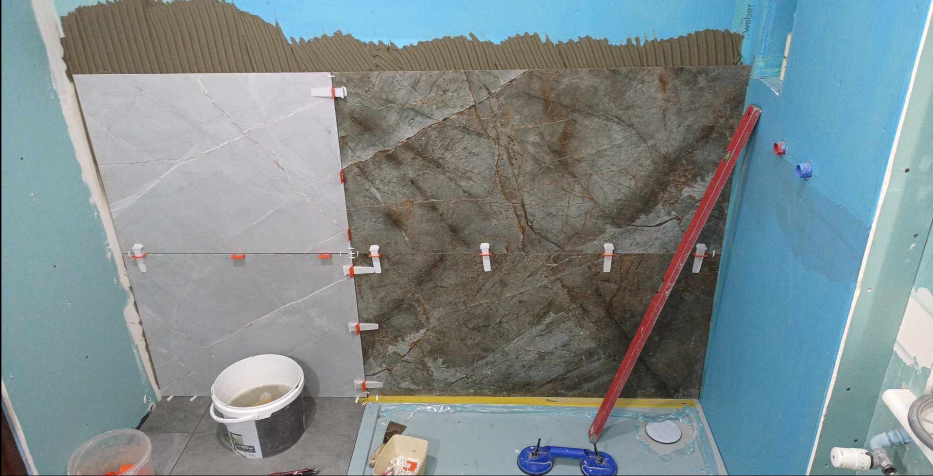 W trakcie układania płytek imitujących marmur w łazience, widoczne poziomica, przyssawka do płytek i wiadro z wodą.