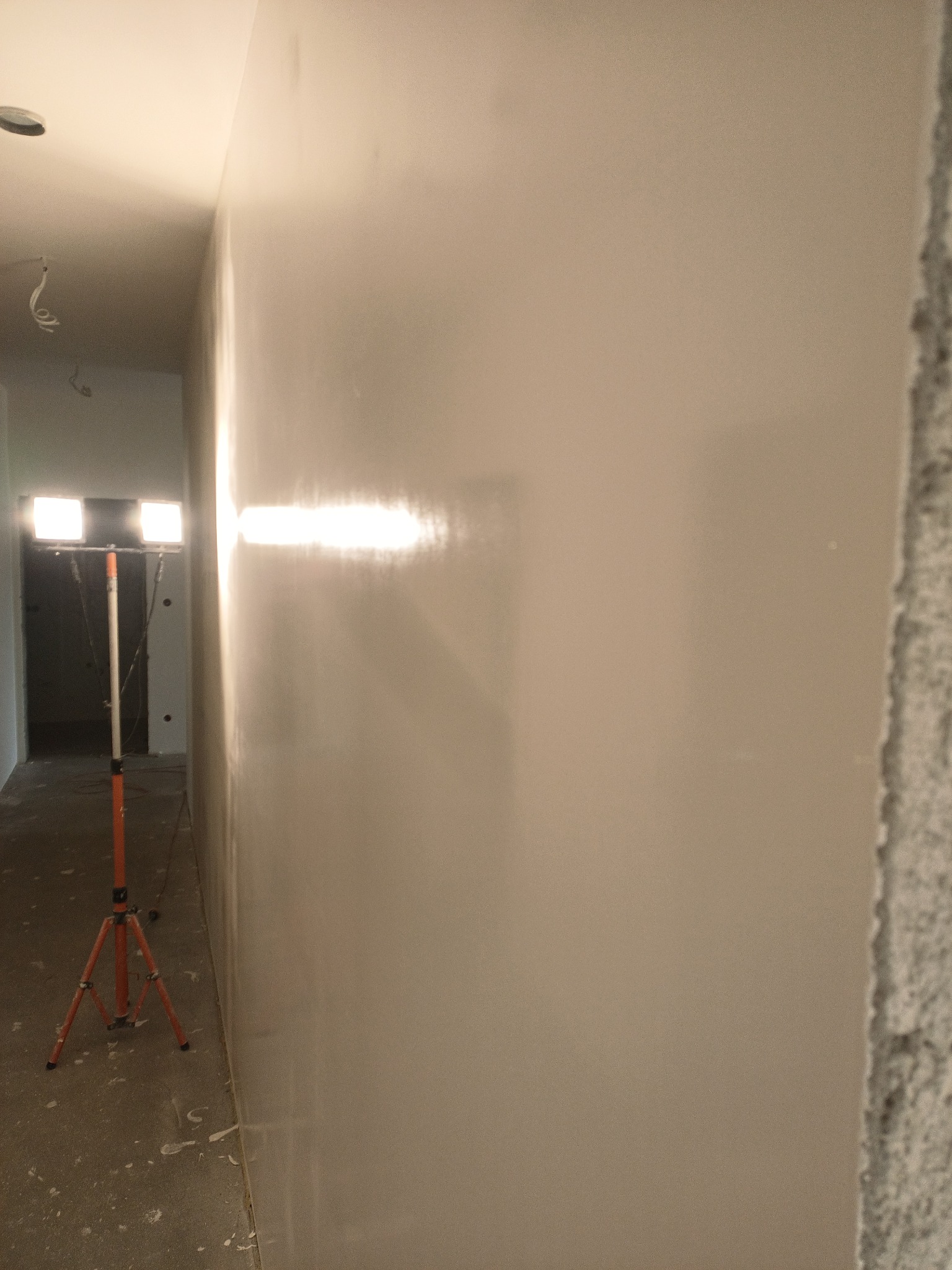 Świeżo nałożona, gładka ściana oświetlona lampą roboczą na trójnogu; widoczny surowy narożnik ściany i przewody pod sufitem.