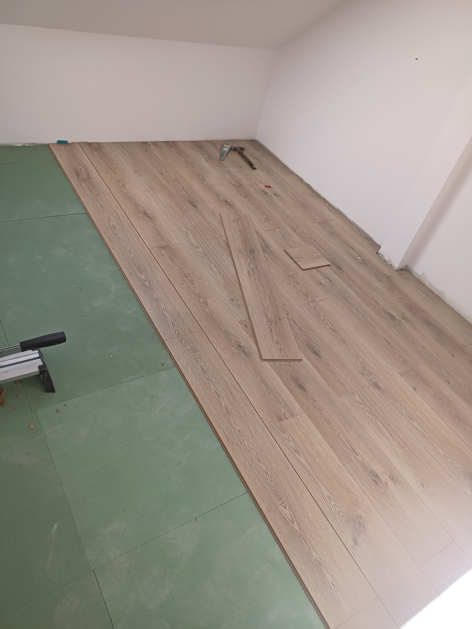 Proces układania paneli podłogowych w pomieszczeniu, widoczna zielona podkładka izolacyjna, częściowo ułożone panele imitujące drewno, narzędzia do cięcia paneli i młotek w tle.