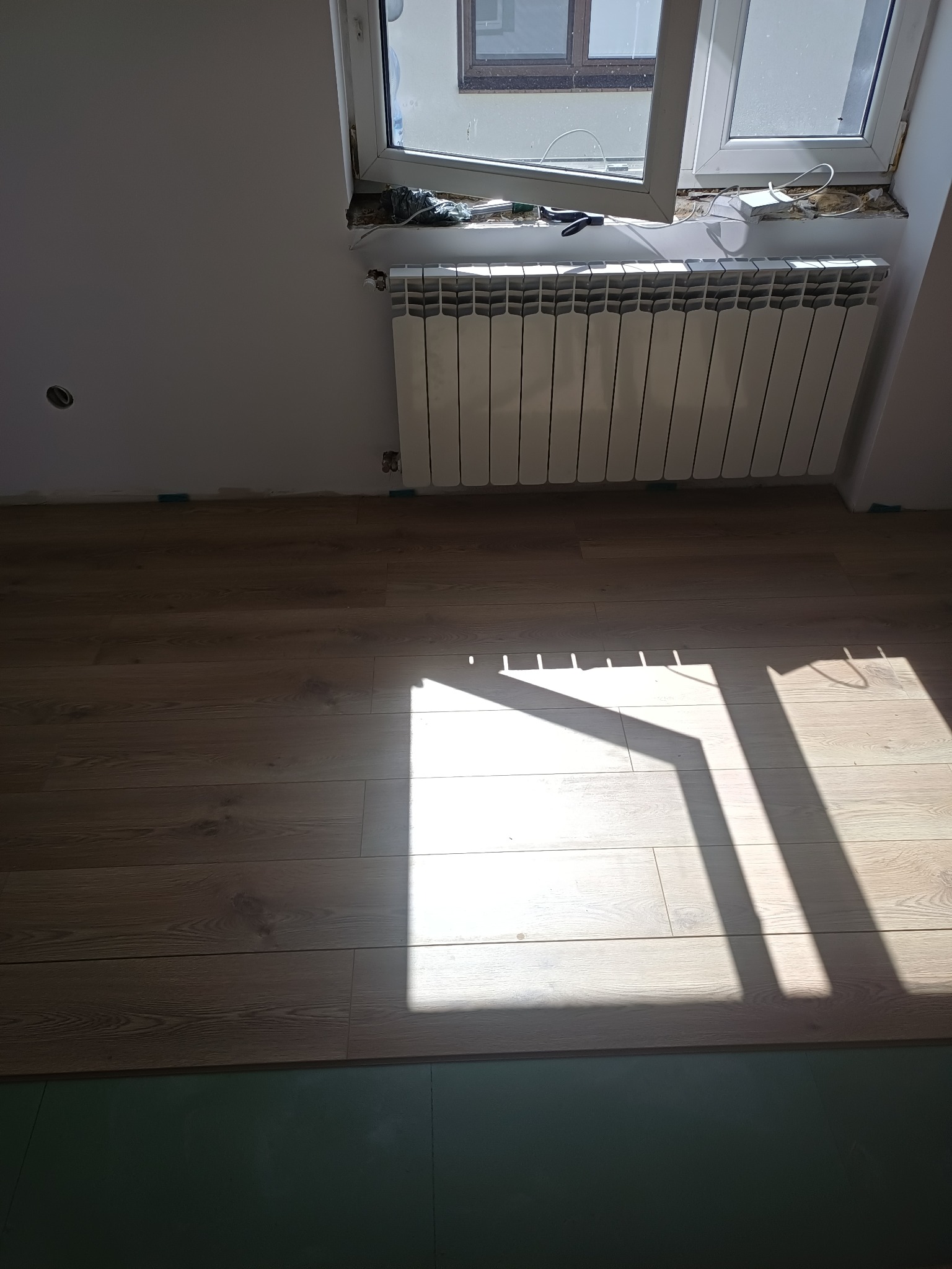 Fragment pokoju z nowo ułożonymi panelami podłogowymi w odcieniach brązu, widoczny grzejnik pod oknem i intensywne promienie słoneczne padające na podłogę.