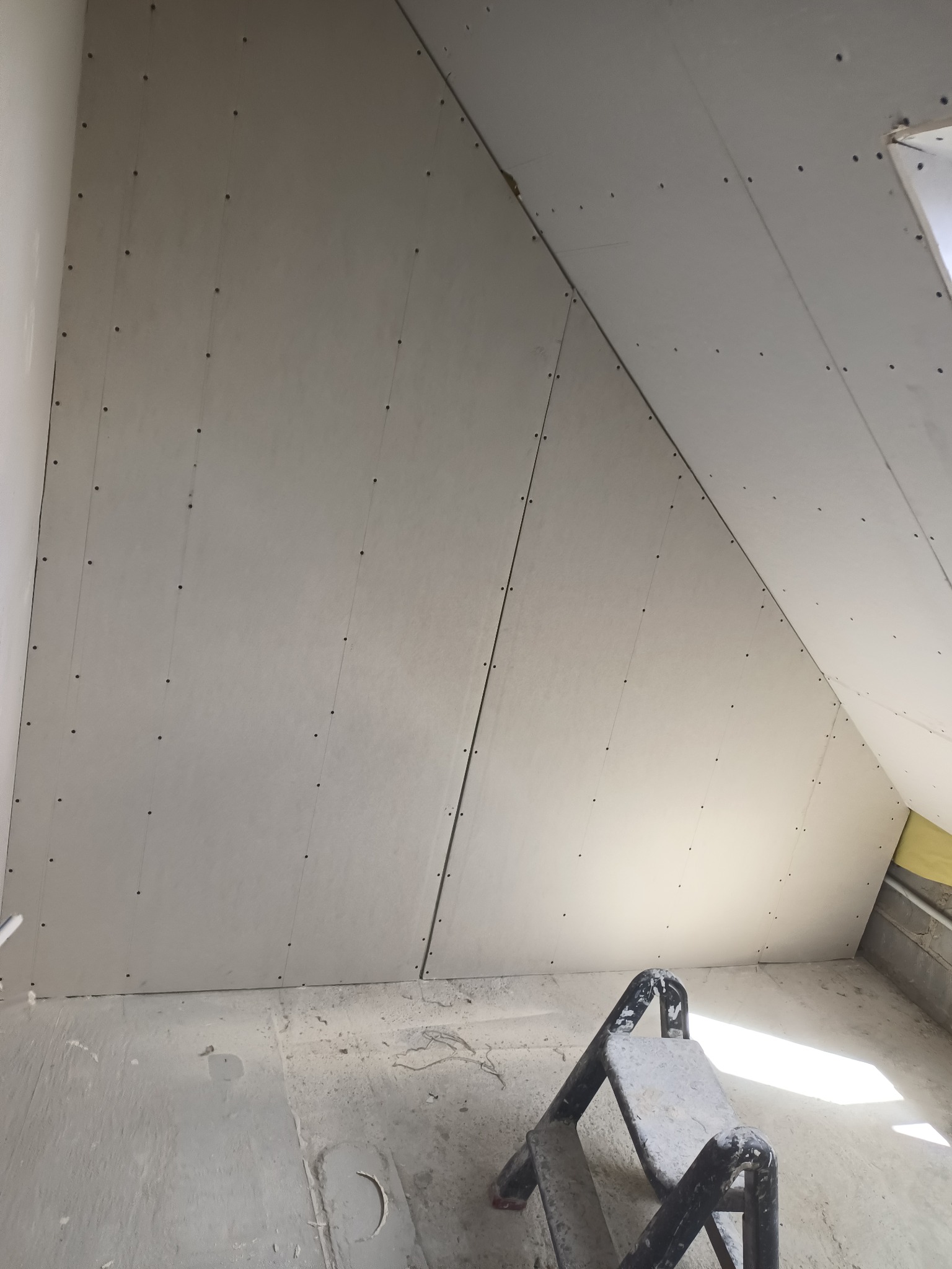 Poddasze w trakcie prac wykończeniowych: płyty gipsowo-kartonowe zamocowane do skośnego sufitu, widoczne ślady po szpachlowaniu na podłodze i drabina malarska.